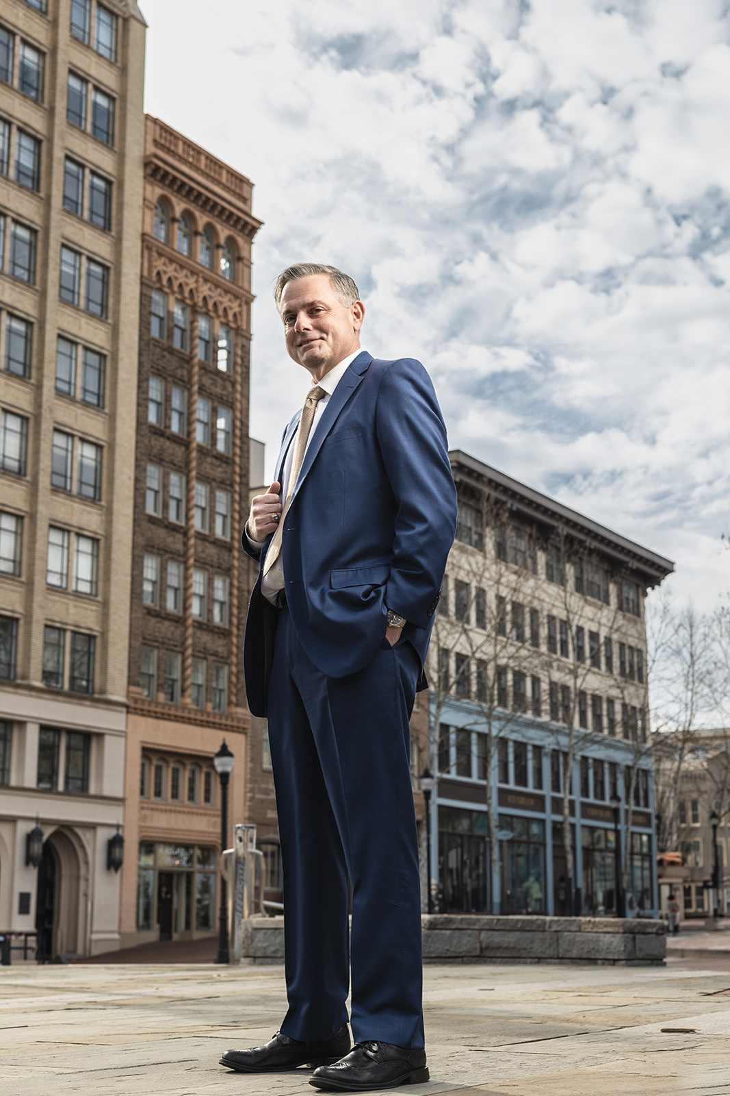 Older White Male in a blue suit standing in front of tall city buildings of Asheville, NC with a cloudy sky background for portrait photography session.