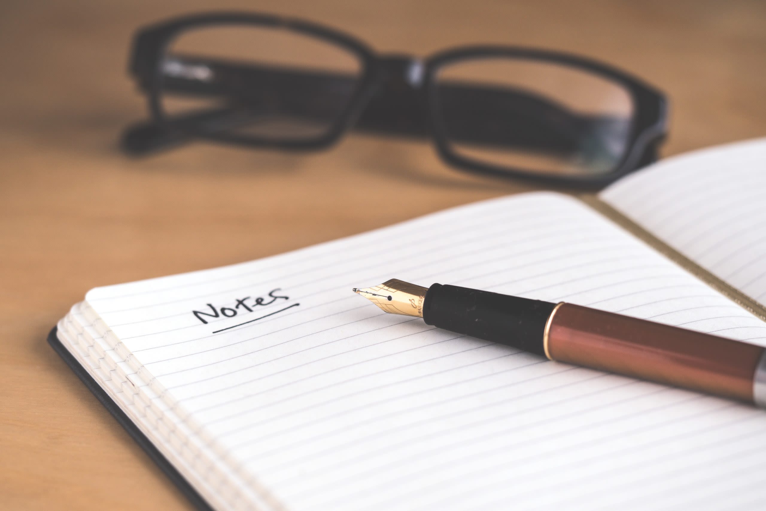 Fountain pen on an open notebook on a desk with eyeglasses sitting in the background