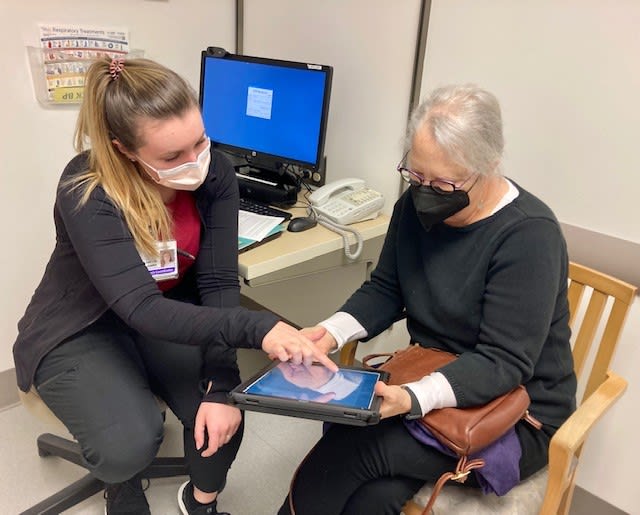 Two women sitting in a medical clinic looking at a tablet. One points to something on the screen as the other observes.
