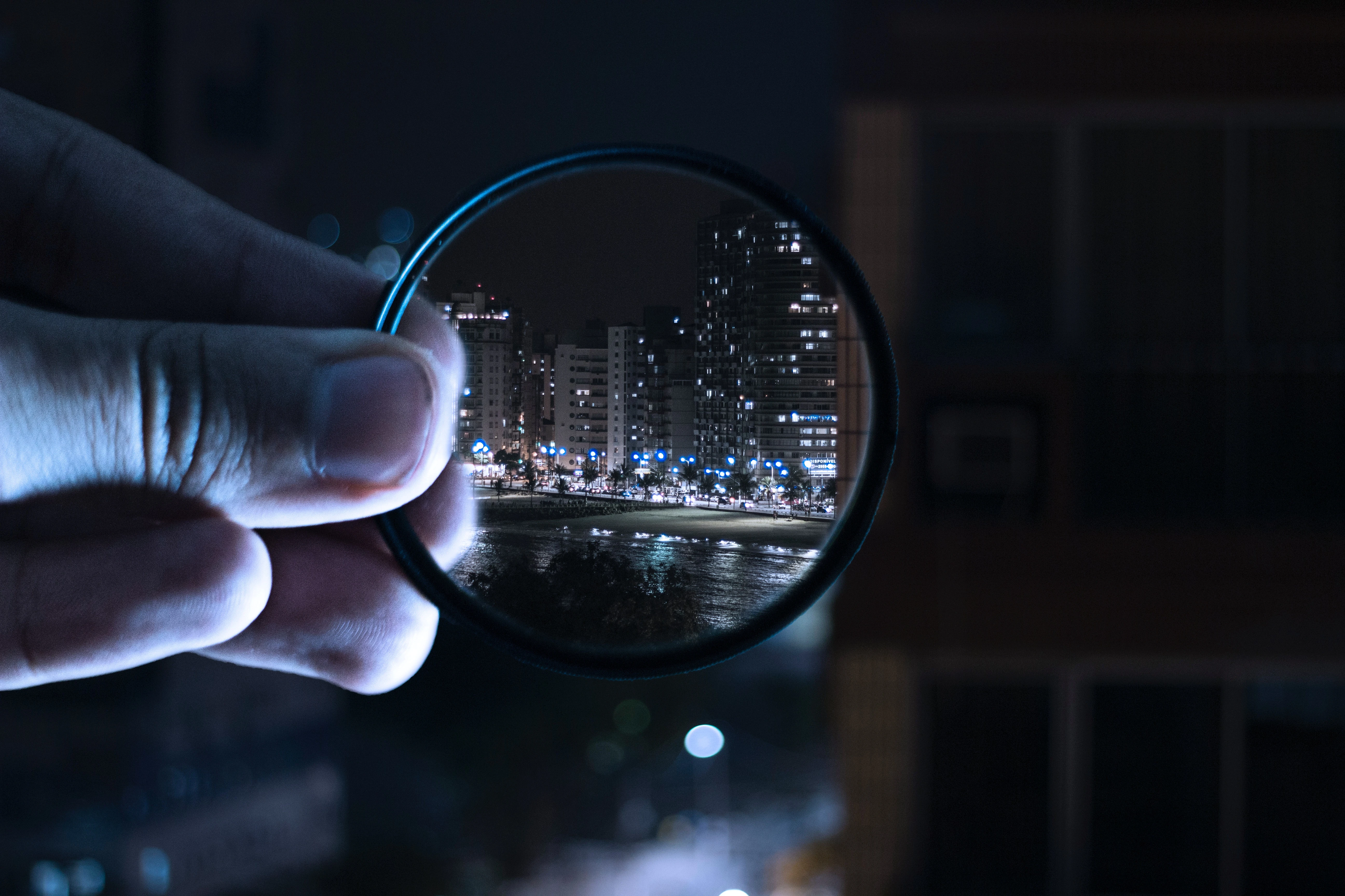 une main approchant un verre de lunette contre une vitre et à l'intérieur duquel on peut voir une image nette des buildings en face