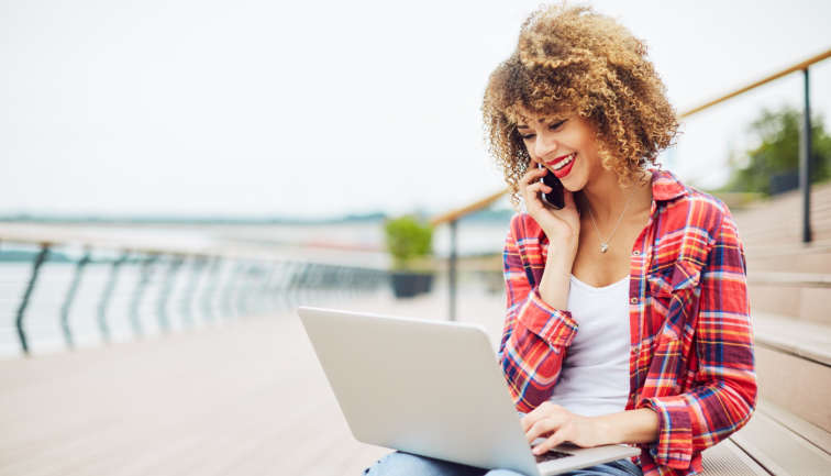 Frau, die mit einem Handy telefoniert und gleichzeitig am Computer schreibt.