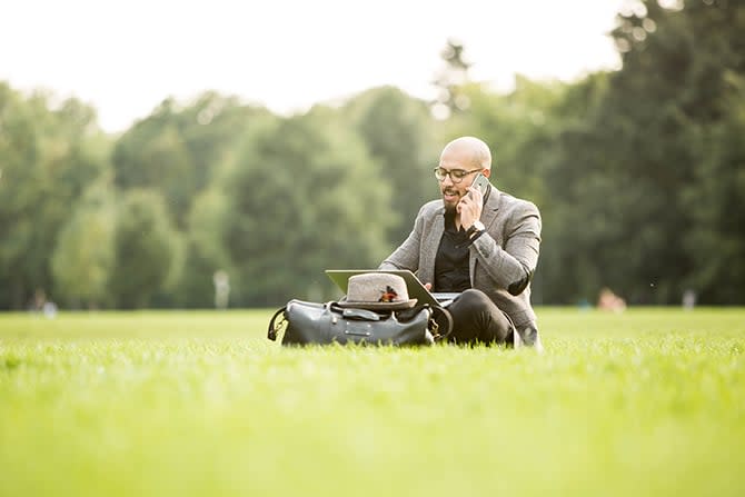 Ein Mann, der in einem Park mit dem Laptop arbeitet und dabei telefoniert.