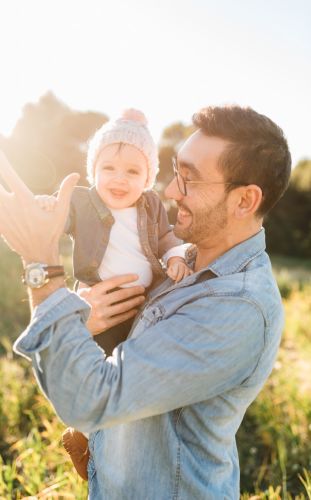 A smiling man holding a baby, standing in a field of tall grass