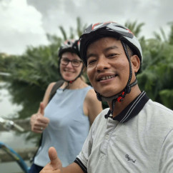 Cyclists show thumbs up next to lush greenery and a river.