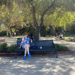 Woman with sunglasses sitting on a bench in a tree-filled area.