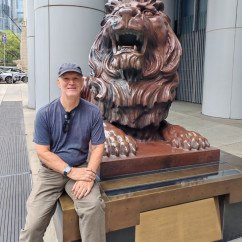 Man seated beside lion statue outside HSBC Main Building, Hong Kong.