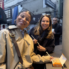 Two women with chopsticks and a food box in a Tokyo alleyway.