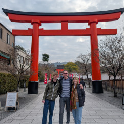 Three people standing in front of a large red torii gate at Fushimi Inari Shrine.