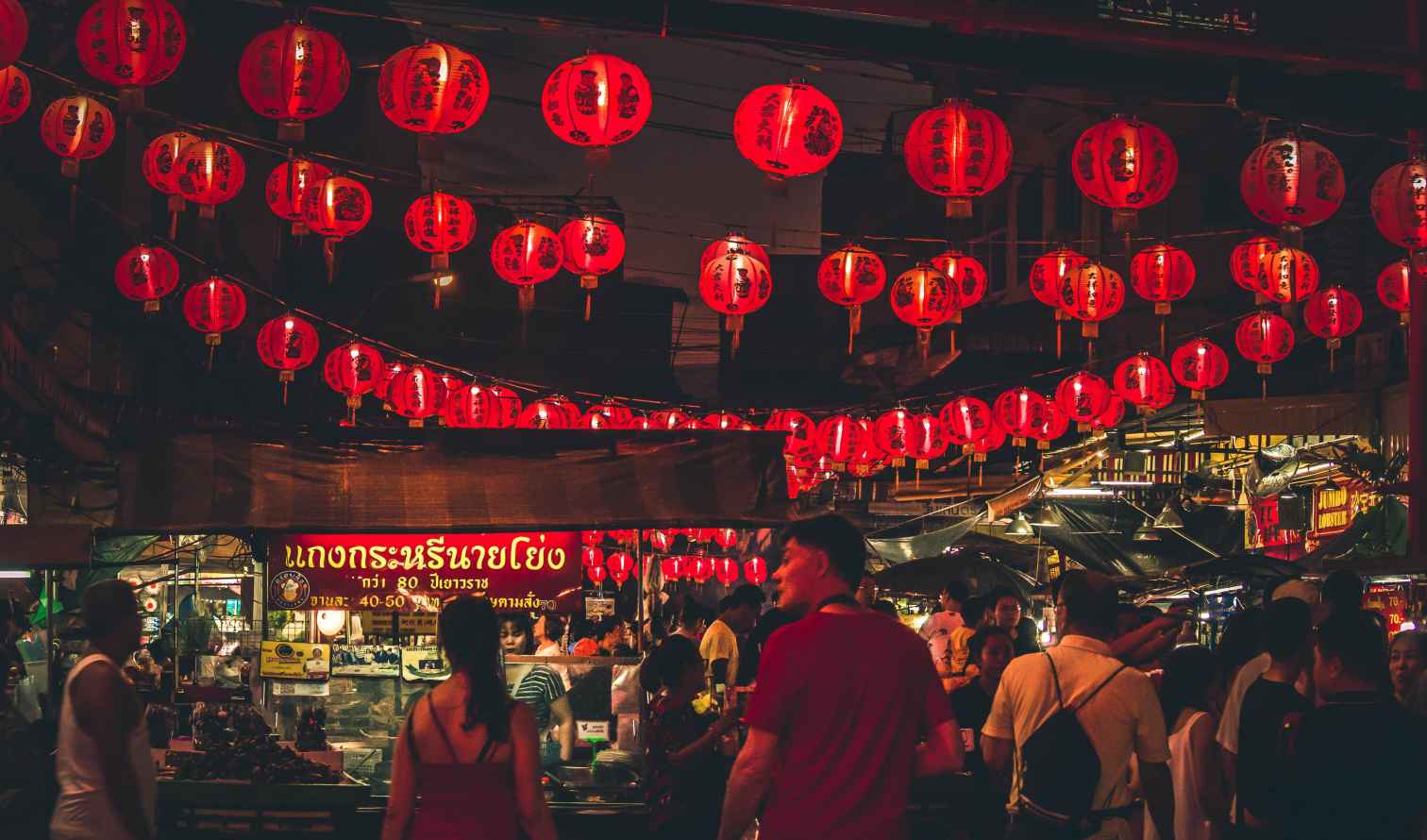 Visitors at a bustling night market under red lanterns in Bangkok