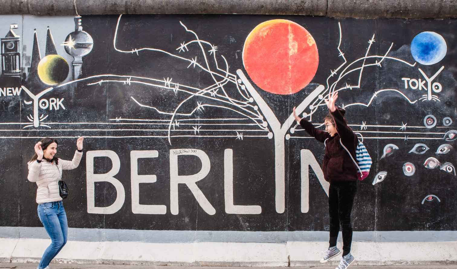Two people pose in front of Berlin Wall graffiti featuring Berlin, New York, and Tokyo.