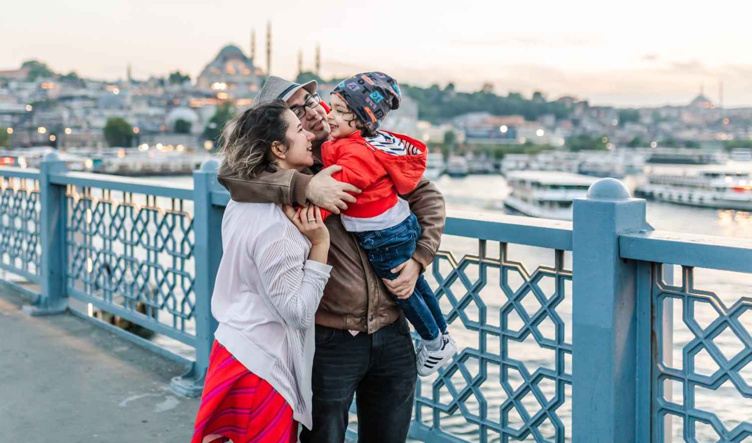 Child in bright jacket held by parents on a bridge in Istanbul.
