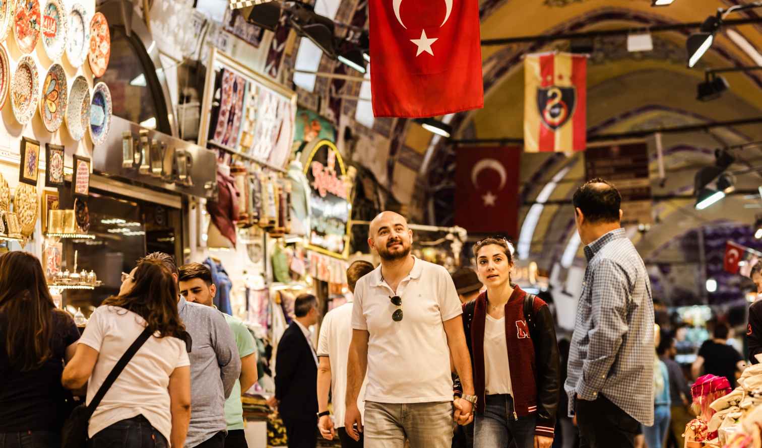 People walking in the Grand Bazaar, Istanbul, under Turkish flags.
