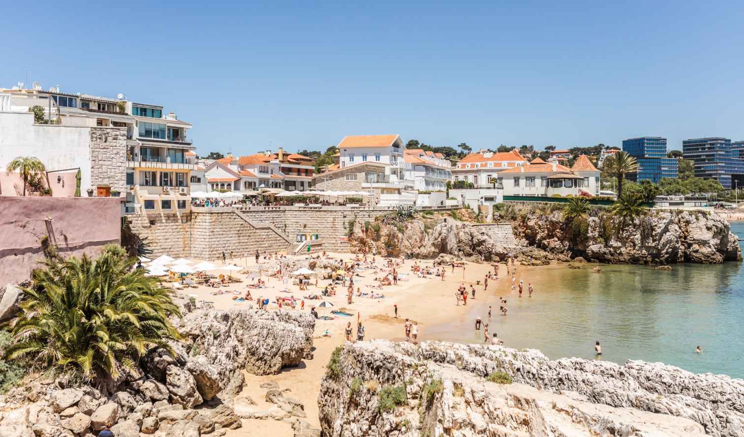 Beachgoers at Praia da Rainha in Cascais, Portugal, with orange-roofed buildings visible.