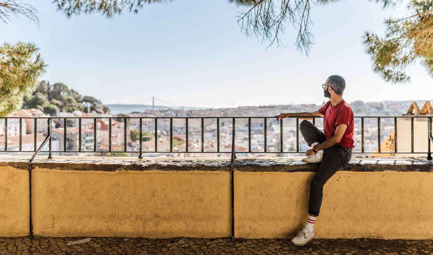 Man sitting on a ledge overlooking Lisbon, Portugal, with the 25 de Abril Bridge visible.