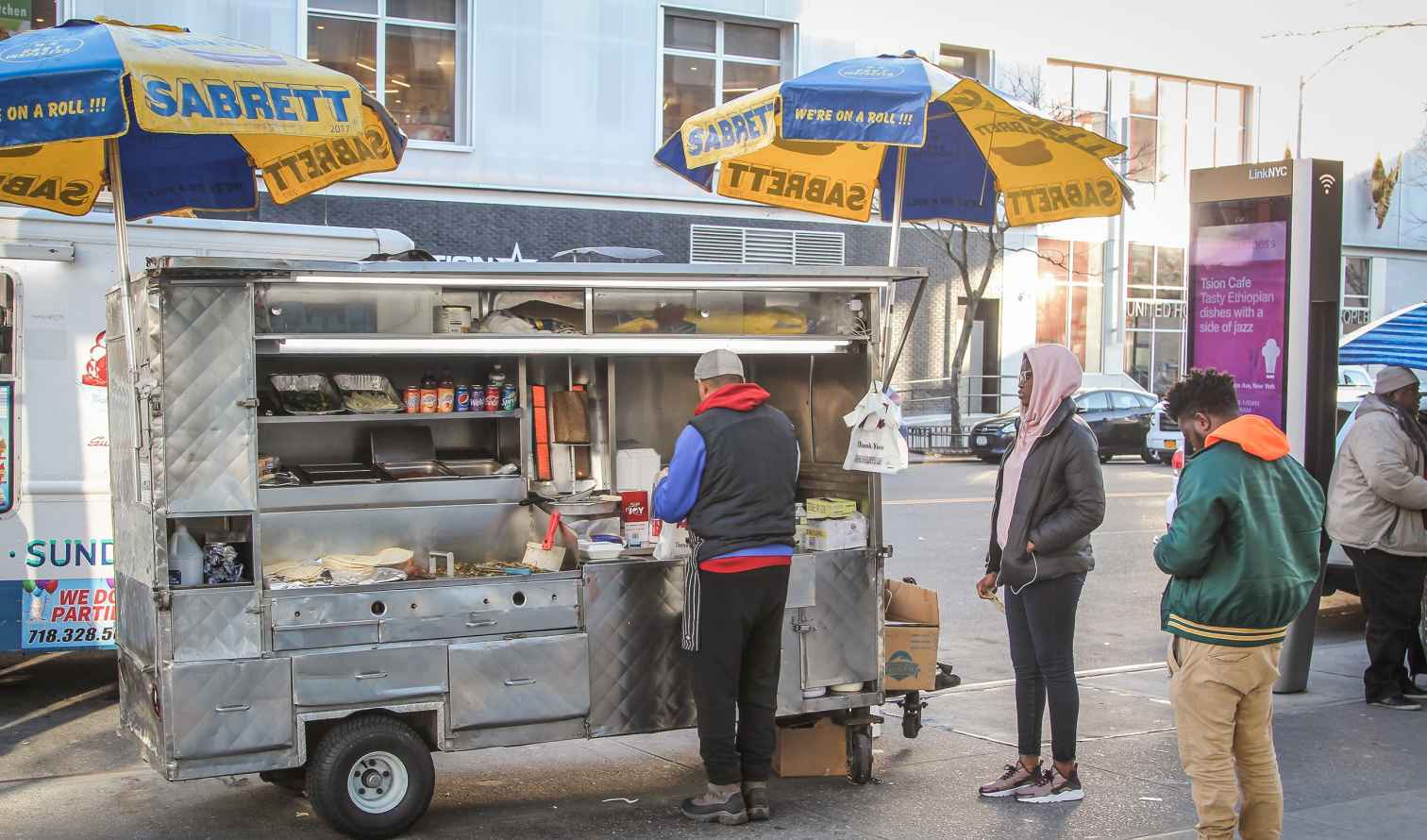 LinkNYC kiosk visible near street food cart and pedestrians.