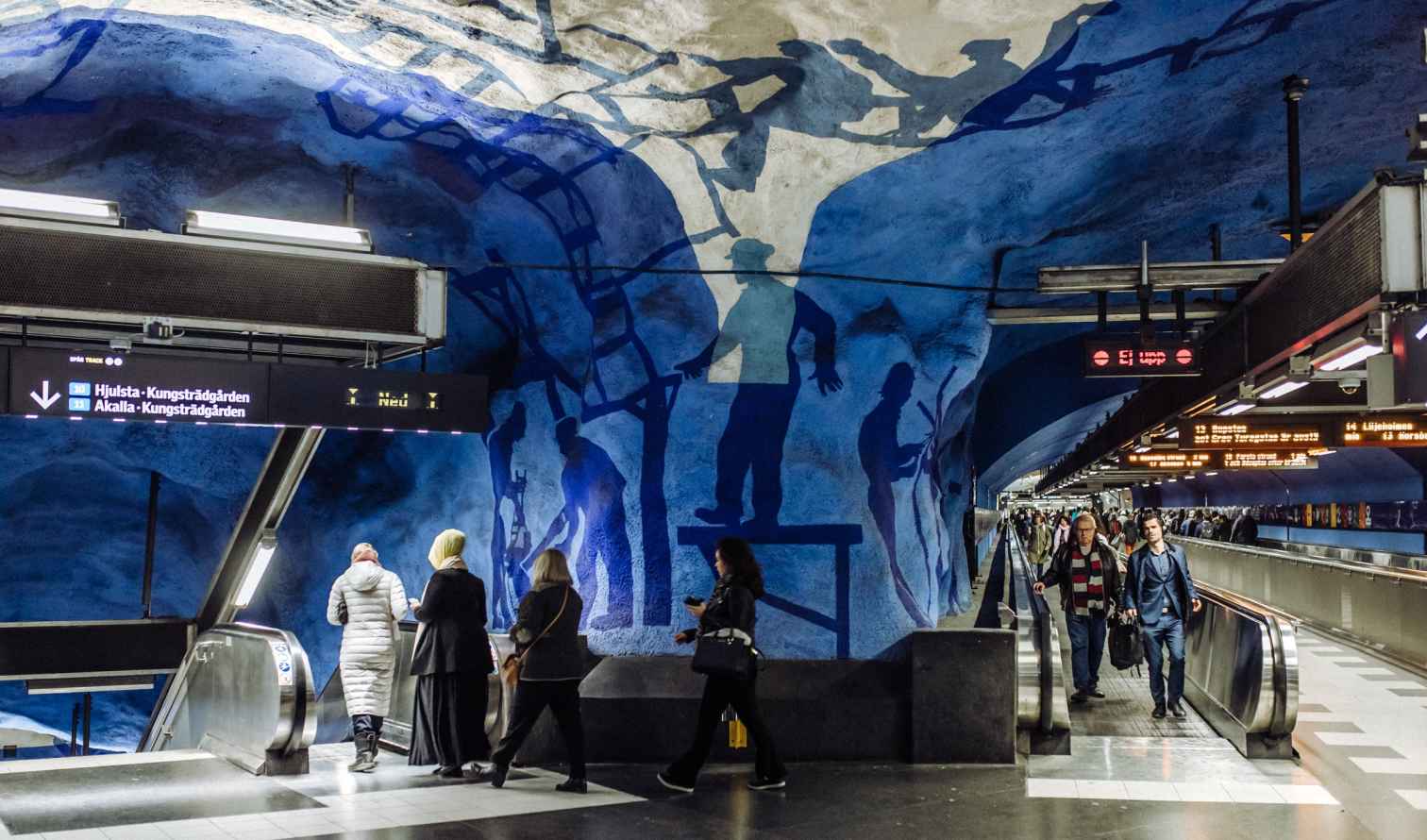 People walking in T-Centralen subway station with blue mural artwork in Stockholm