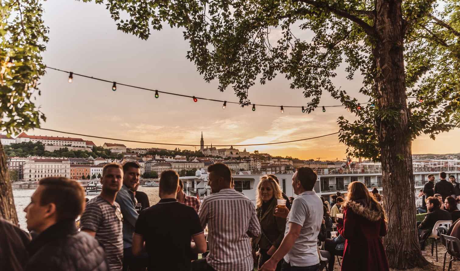 Groups of people by the riverside with Budapest architecture visible.