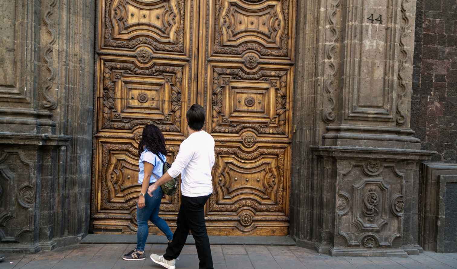 Two people walking past a large ornate wooden door in Mexico City