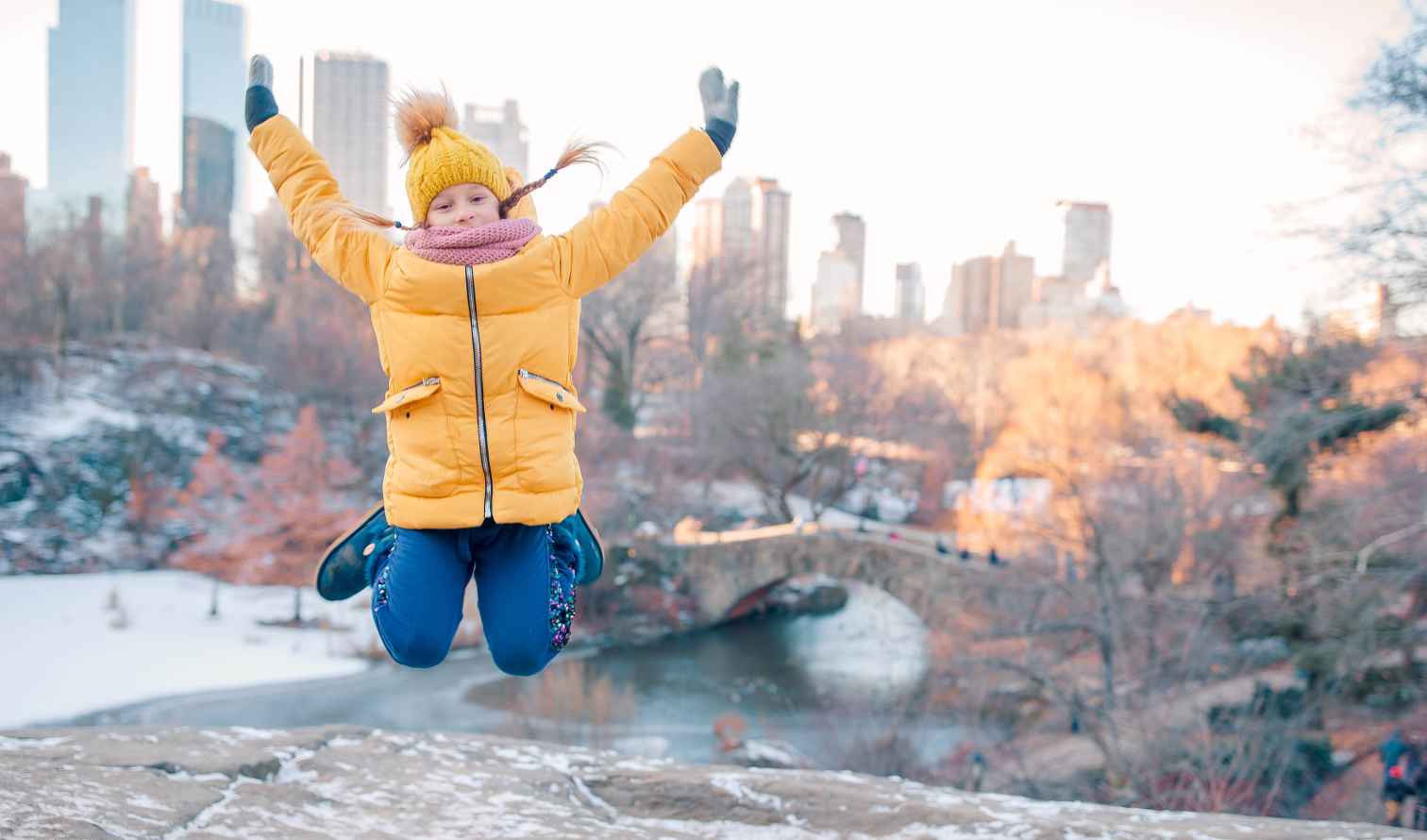 Snow-covered rocks and child jumping, Gapstow Bridge visible behind in Central Park in New York