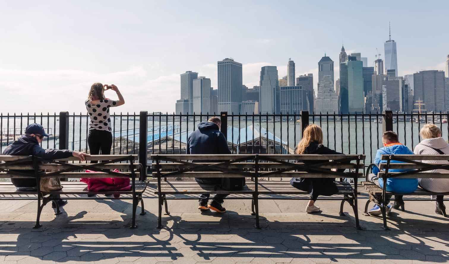 People sitting on benches at Brooklyn Heights Promenade overlooking Manhattan skyline.