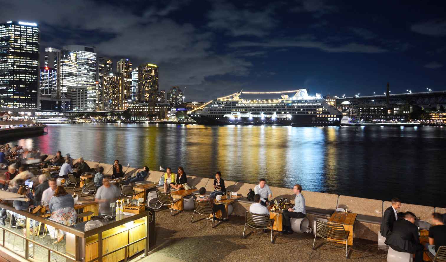 People dining outdoors near Sydney Harbour with city buildings in the background.