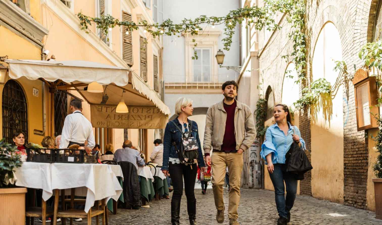 Three people walk past a restaurant on a narrow street in Rome.