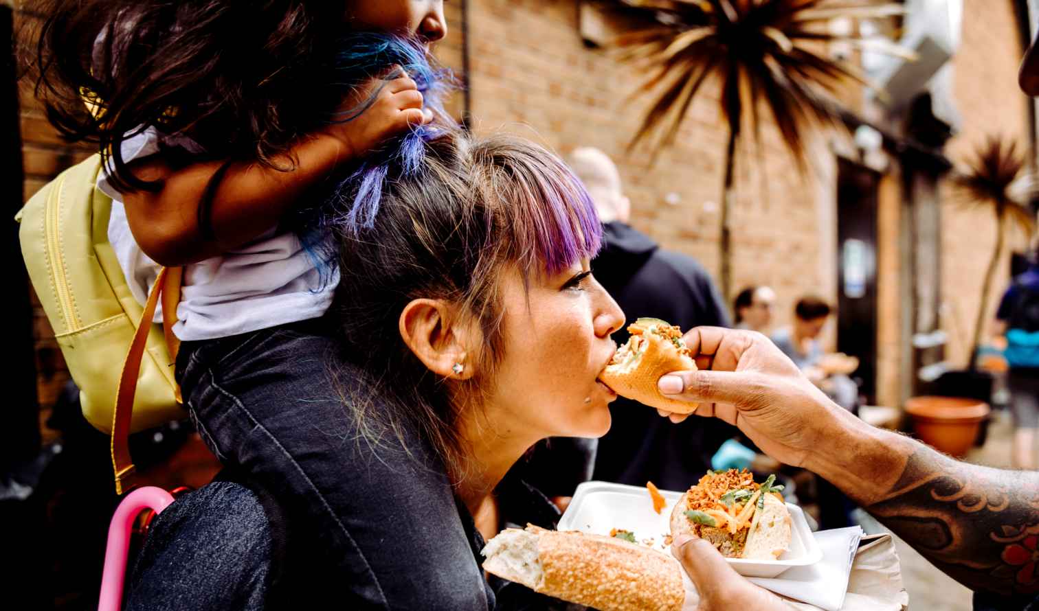 Woman with child on shoulders eating a sandwich on Brick Lane, London.