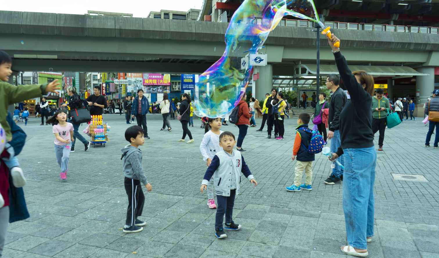 Adults and children gathered in urban square, Taipei Main Station visible.