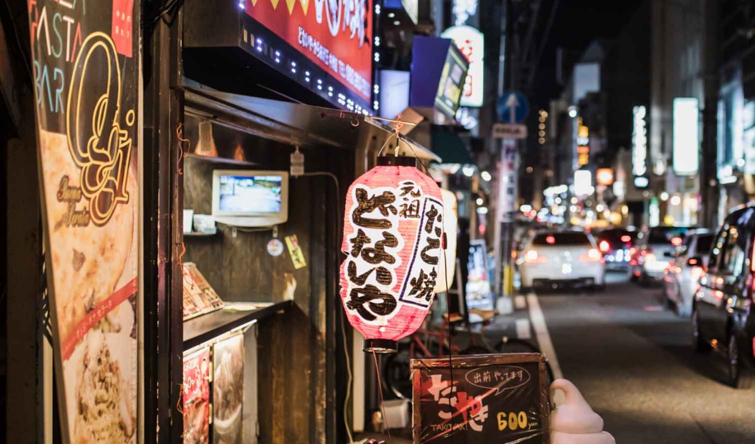 Street view in Fukuoka, Japan, with illuminated signs and passing cars.