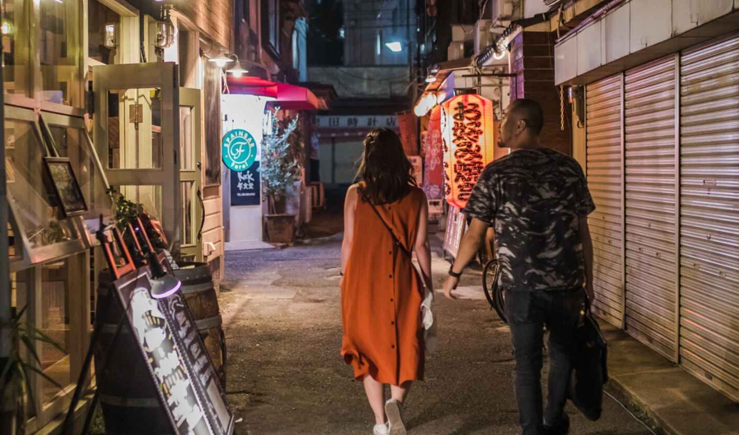 Two people pass by closed shops and lit lanterns in an urban alley in Fukuoka