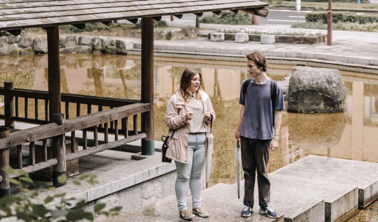 Two people standing near a traditional wooden pavilion by a pond in Nagoya