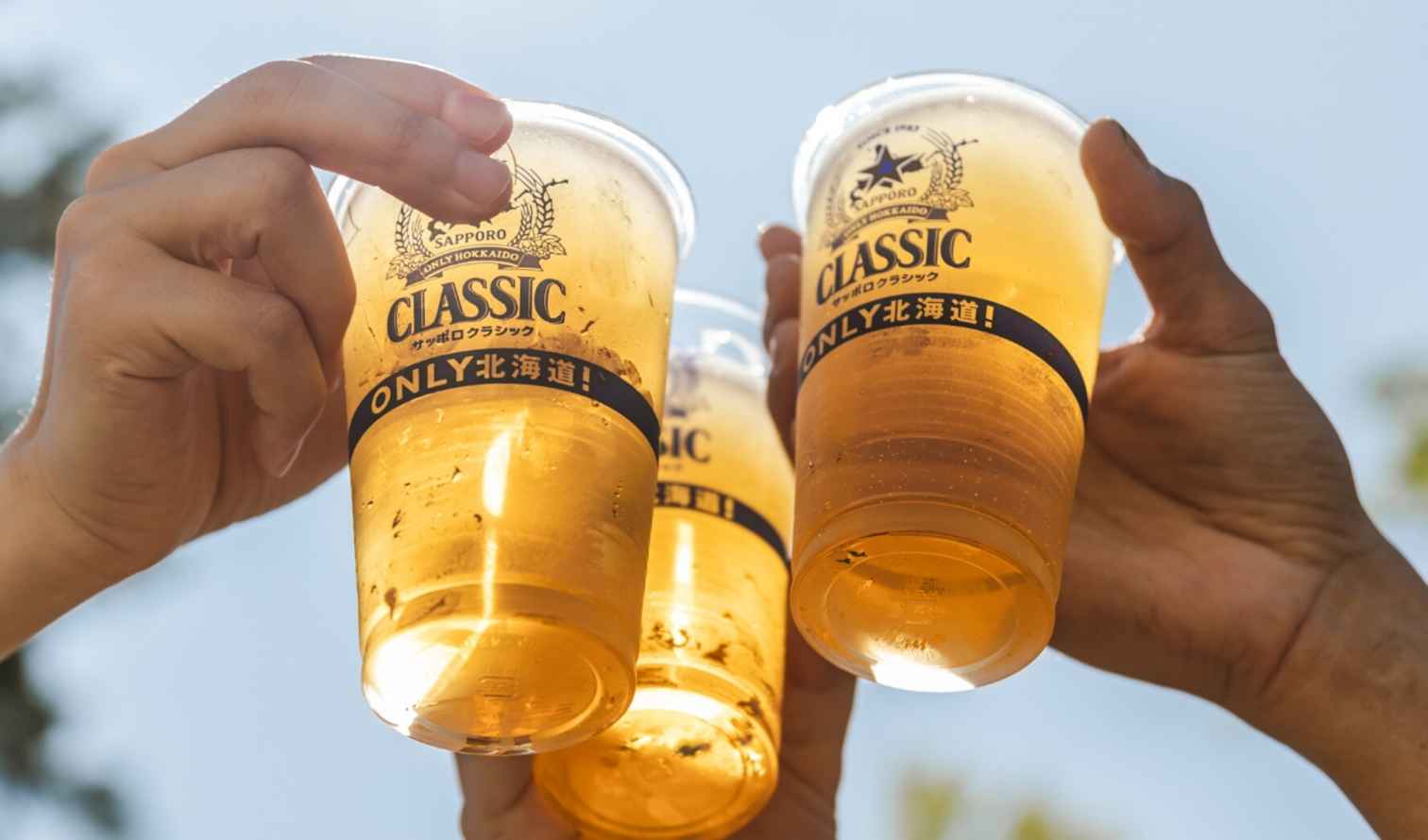 Three hands holding Sapporo Classic beer cups in an outdoor setting.