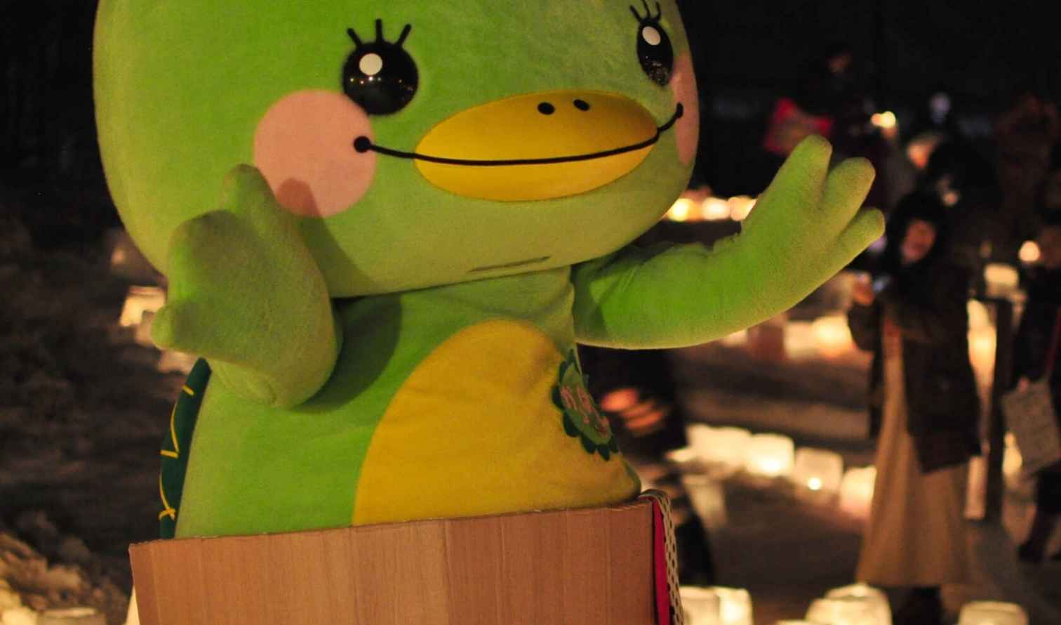 People gather around lanterns in the snow next to a giant green mascot character.