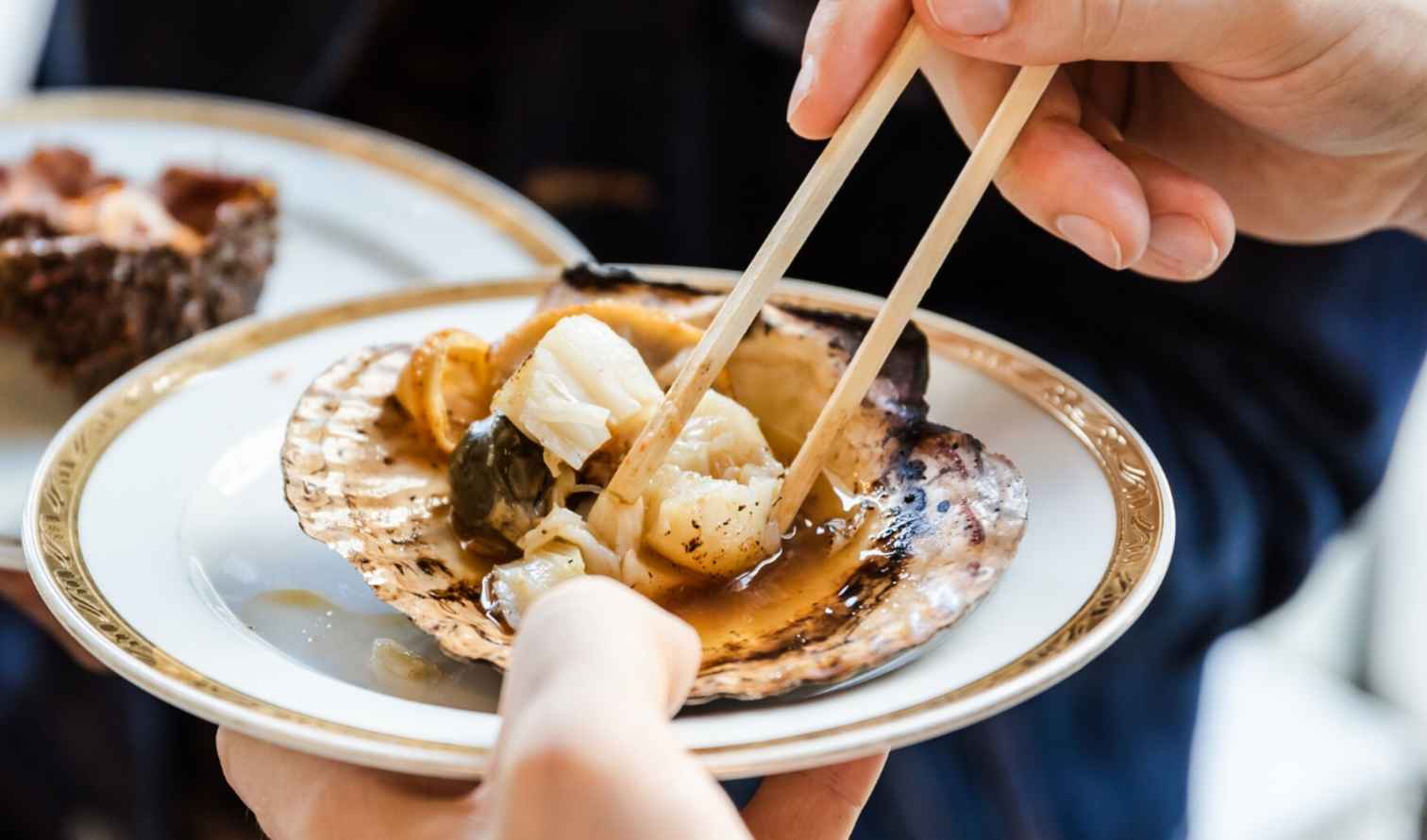 Person using chopsticks to eat scallops from a decorative plate in Sapporo