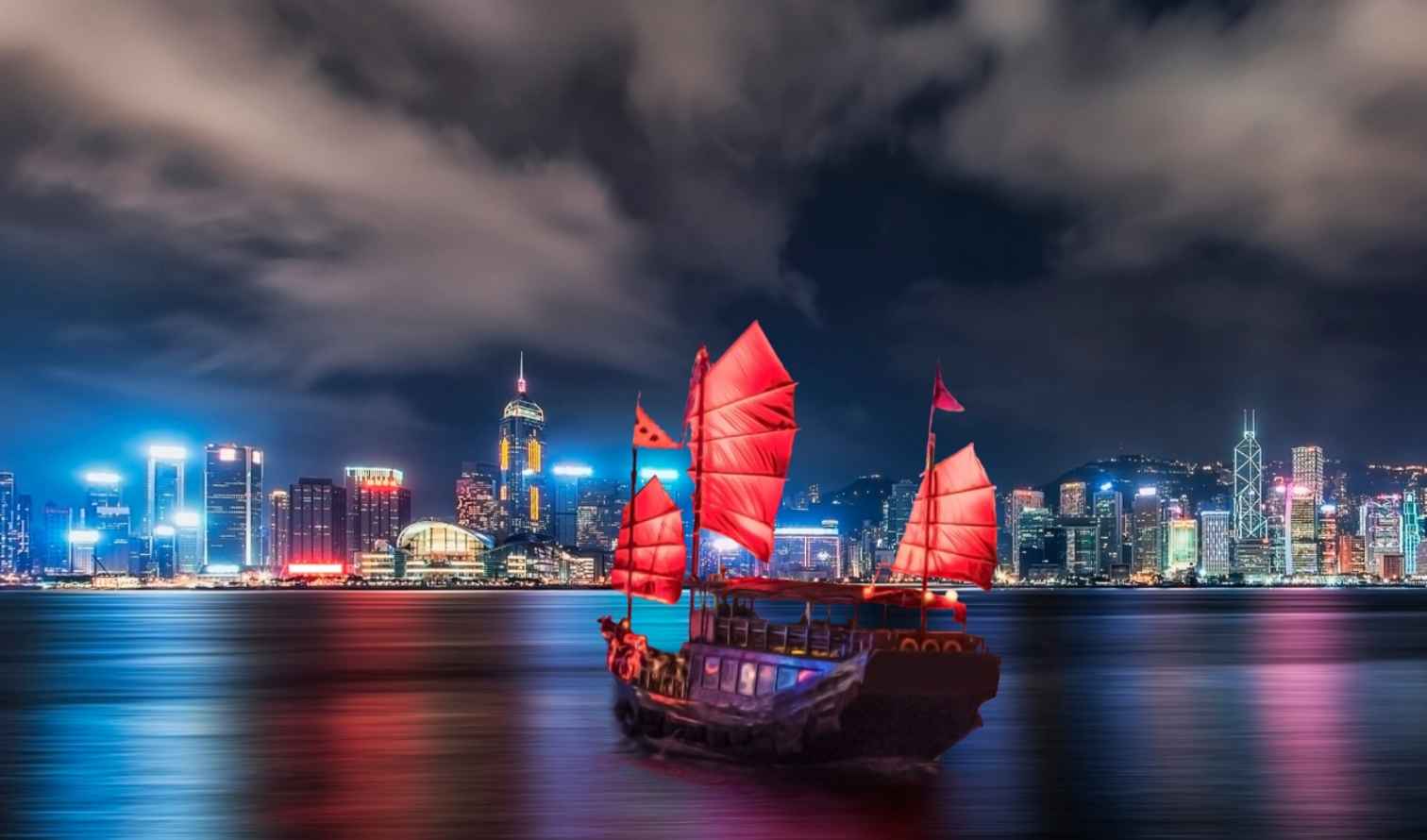 Traditional junk boat on Victoria Harbour with Hong Kong skyline at night.