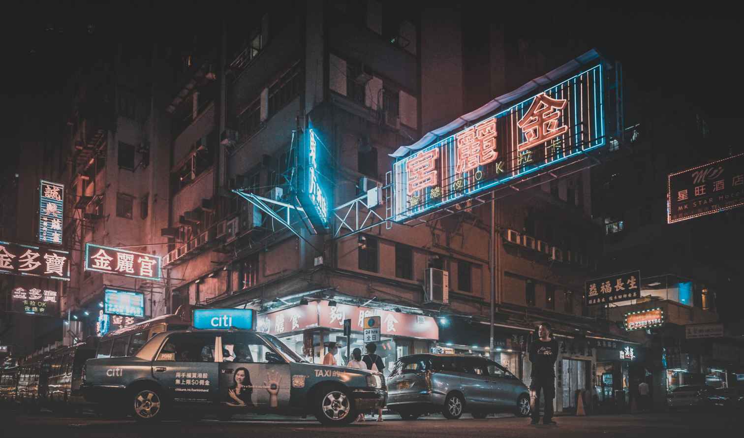 Nighttime street scene in Hong Kong with neon signs and parked cars.