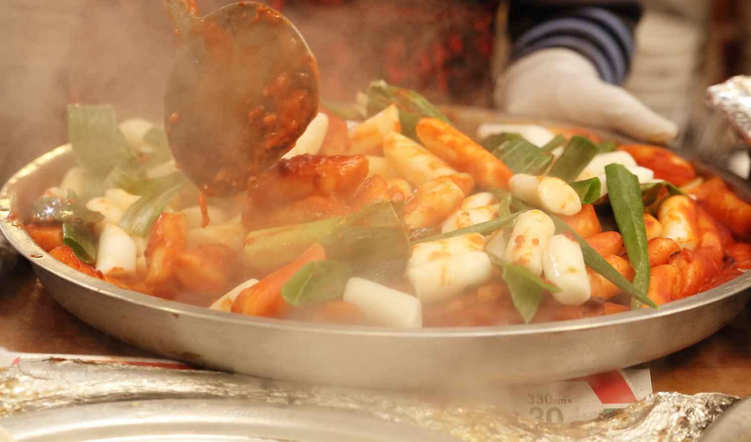 Close-up of a hand with a glove ladling sauce over tteokbokki in Seoul