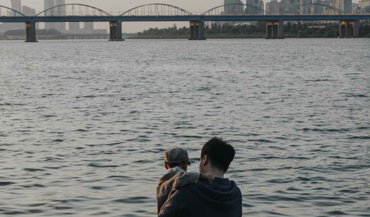 Man holding child by Han River with bridge and city skyline in Seoul.