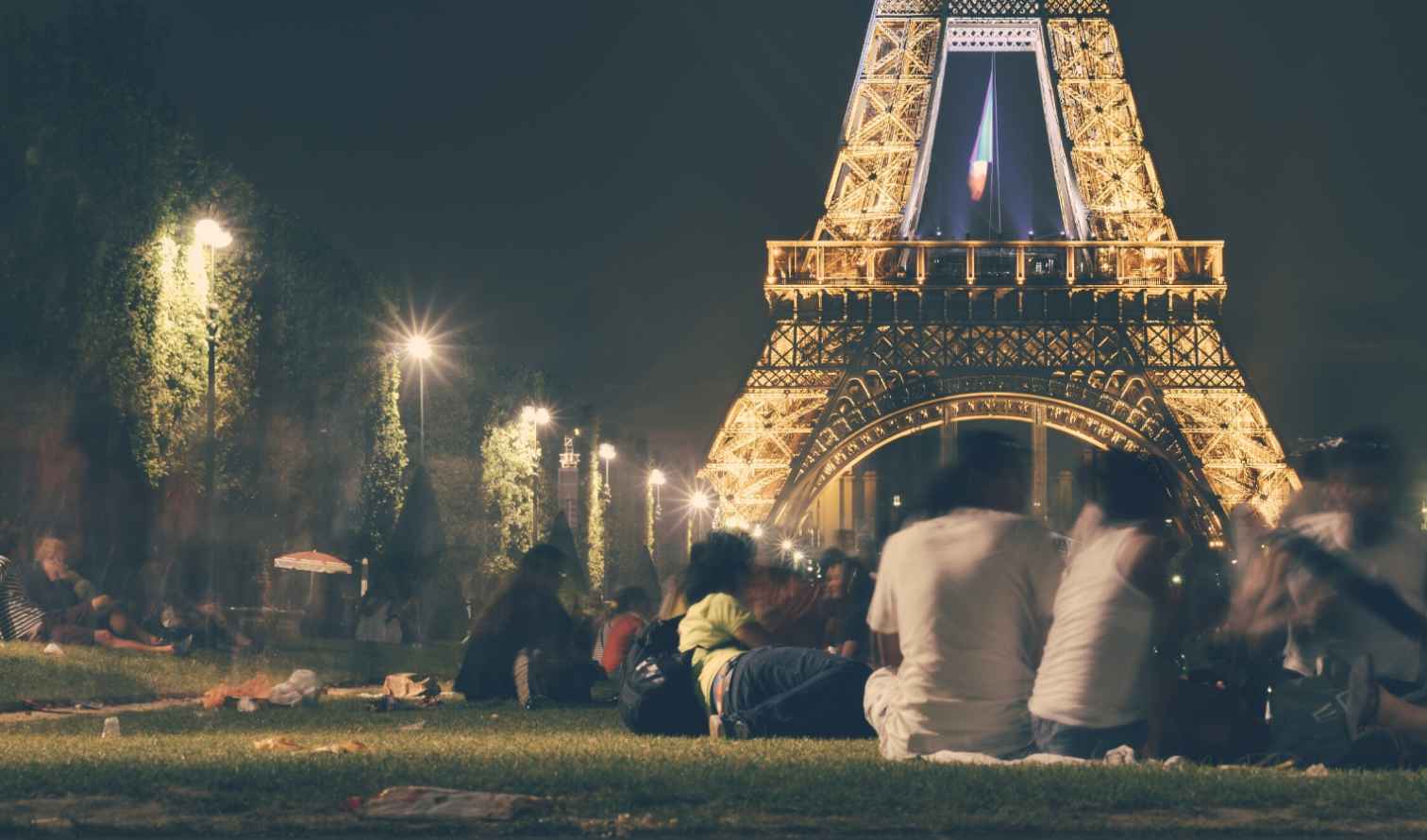 People sitting on grass near the illuminated Eiffel Tower at night.