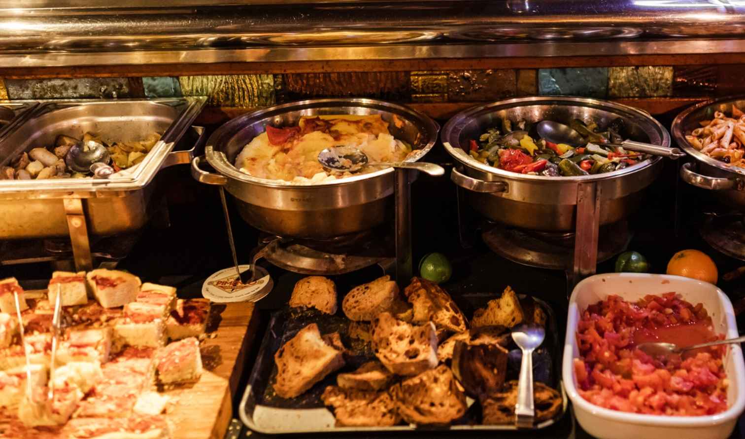 Buffet table with trays of assorted dishes, including pasta and roasted vegetables in Milan