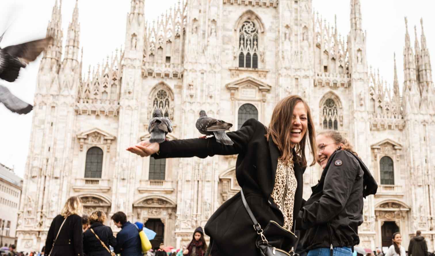 Two women smiling in front of Milan Cathedral with pigeons on their arms.