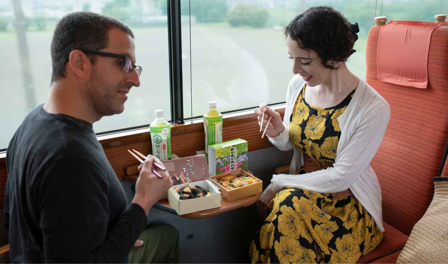 Two people eating bento meals on a train with large windows in Tokyo
