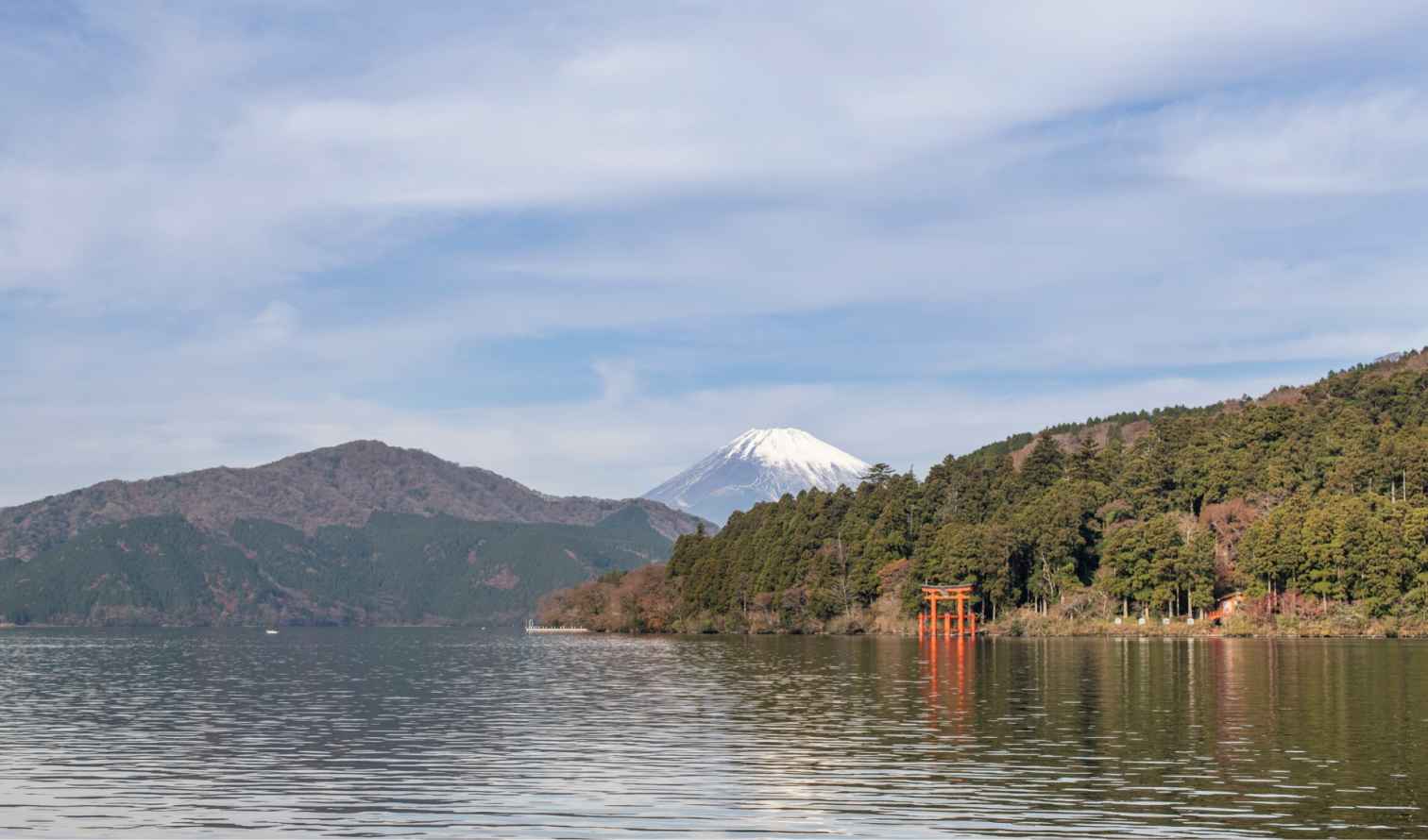 Snow-capped Mount Fuji viewed from Lake Ashi, bordered by forest.