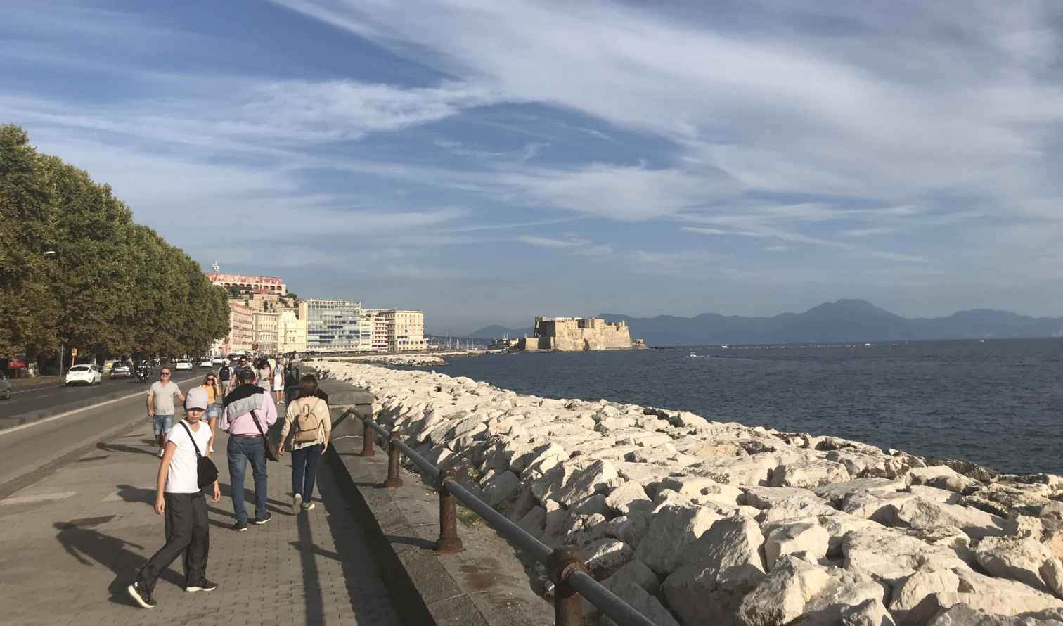People walking along Via Partenope in Naples, with Castel dell'Ovo visible.