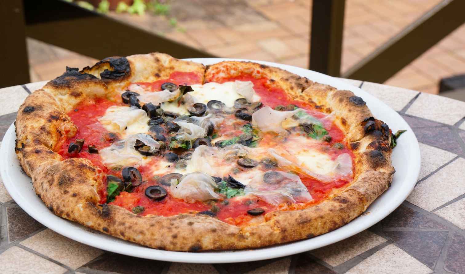 Wood-fired pizza with olives, cheese, and prosciutto on a patio table in Naples