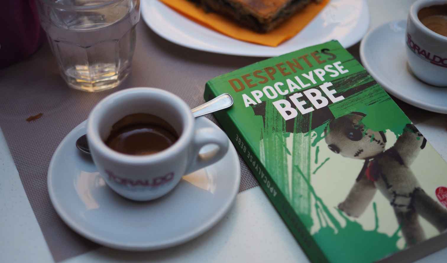 Close-up of a book titled 'Apocalypse Bebe' beside a cup of coffee in Naples