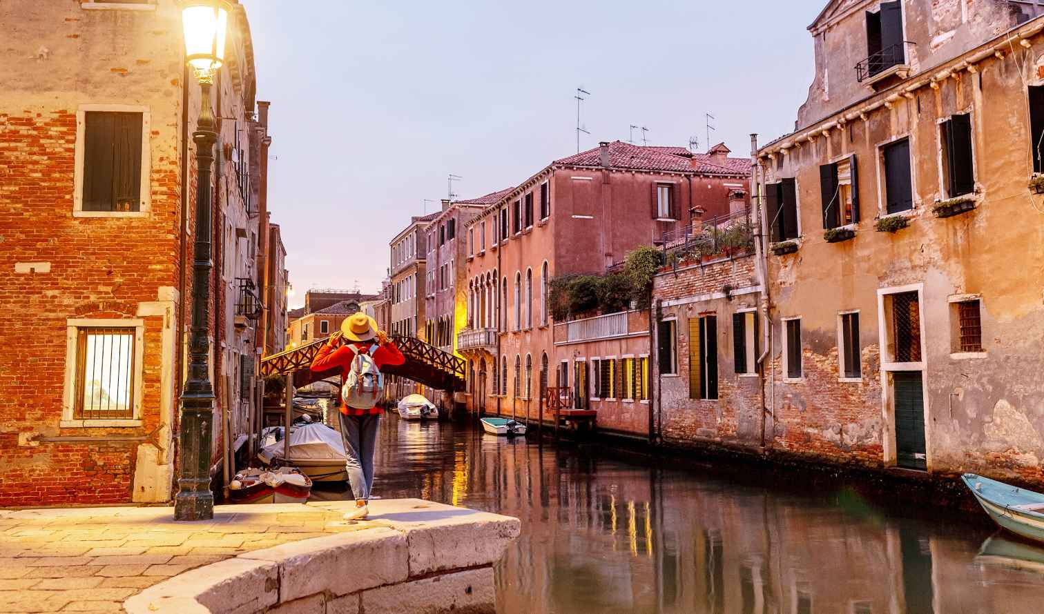 A person stands beside a canal in Venice near a lit street lamp.