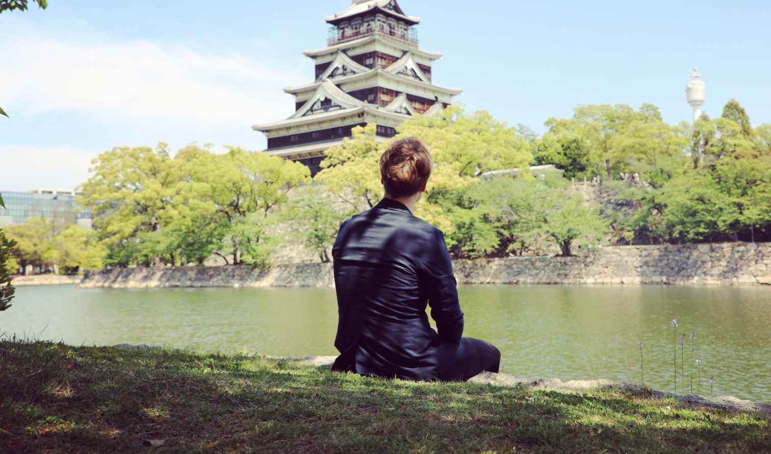 Person sitting by the water, looking at Hiroshima Castle with trees in view.