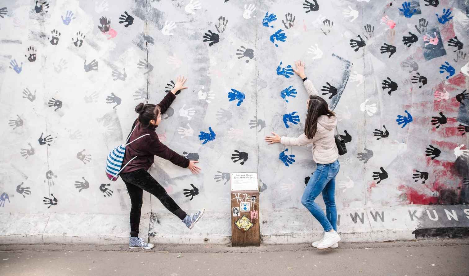 Two people interact with handprint art on the Berlin Wall.