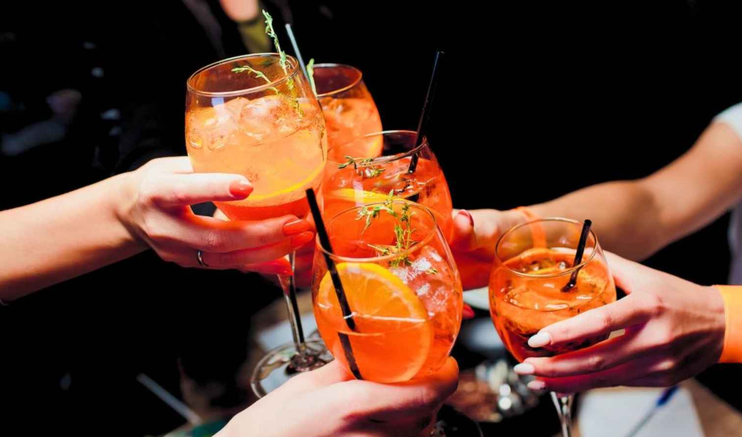 Five hands holding glasses of orange cocktails with straws and garnishes in Milan